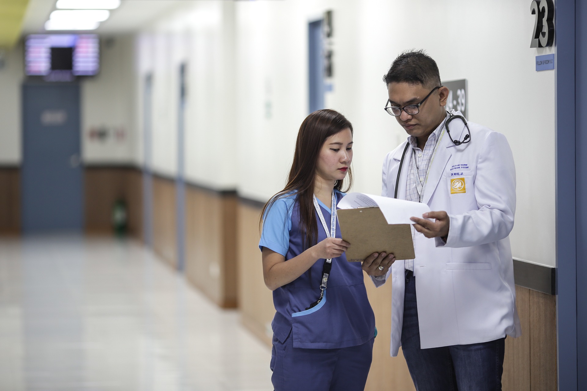 Male doctor in white lab coat sharing medical record with nurse in blue scrubs in clinic hallway for clinical start-up.