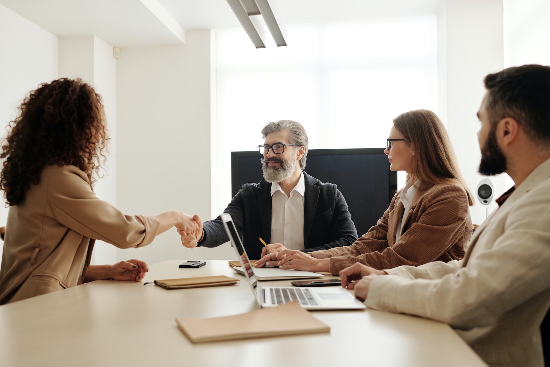 Four people in business attire seated around wooden table shaking hands for clinical research recruitment (HR) meeting.